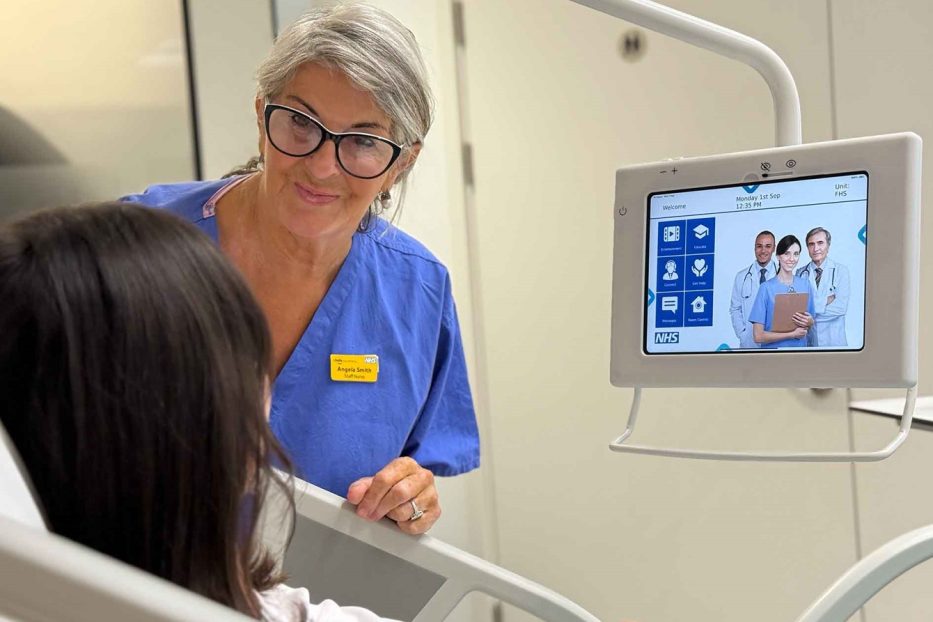 NHS nurse supporting patient beside digital bedside screen in a future healthspaces hospital room.